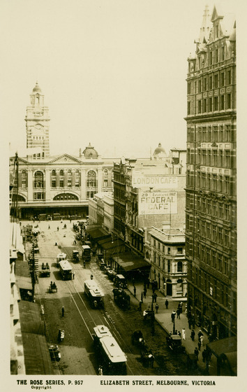 Photograph taken from a tall building, looking down at a streetscape. Tall buildings stand on either side of the street and trams are lined up in the middle of the road. At the end of the street, the facade of Flinders Street Station, including the clock tower, can be seen.