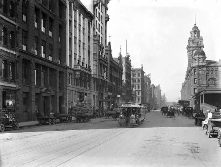 Streetscape of Melbourne in the early 1900s. Tall sandstone or brick buildings stand either side of the street. A tram carrying passengers is in the middle of the street. Further in the distance commuter traffic of cars and horse carriages can be seen.