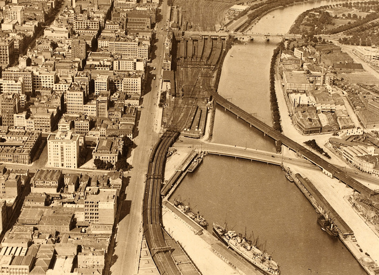 Aerial view of a large cityscape. Through the middle of the picture is a large river and a series of trainlines running adjacent. On either side of the river are lots of city buildings.