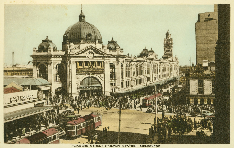Postcard of a streetscape in Melbourne. The main building in the postcard is Flinders Street Station, showcasing its facade and size compared to nearby buildings. In the centre of the street, trams, cars and pedestrians walk past the station and the nearby buildings.