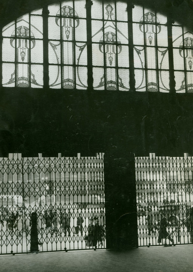 From inside the station looking back towards the entrance way which is closed off by metal gates. Above the gates is a large arched window filled with stain glass and patterns.