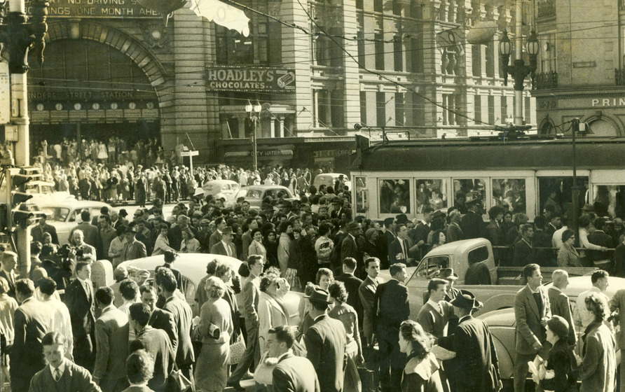 A large crowd of commuters walk past the entrance way to the train station. They are intermingled with cars and trams as they walk on the road.