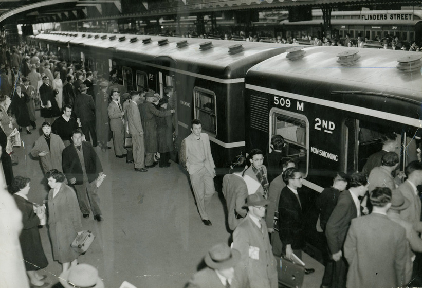 A large group of people stand on a train platform. Some people are getting onto the train, whilst others stand around and talk.