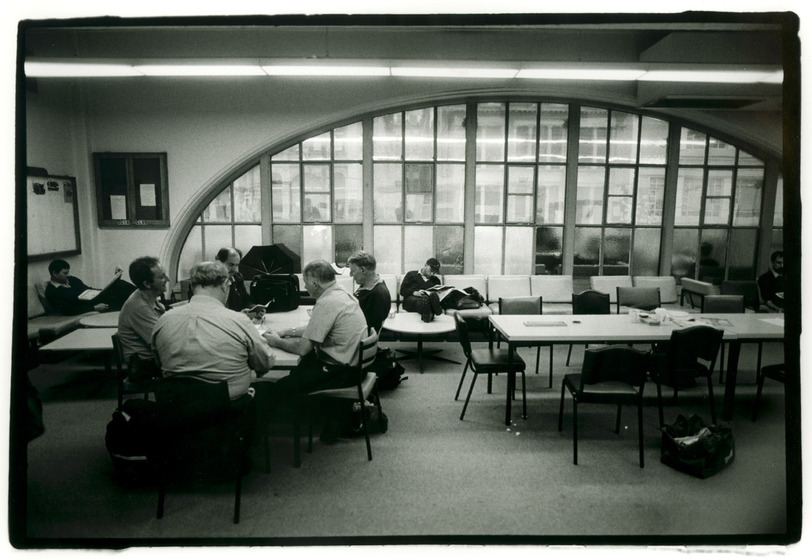 The inside of a room with people sitting around tables talking, whilst others sit solo and appear to be reading. Behind the tables is a large glass paneled arched window.