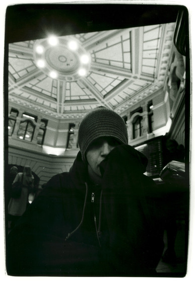 A young man in a beanie stares down into the camera. Behind him the decorative domed roof of the inside of the Flinders Street Station entrance can be seen.
