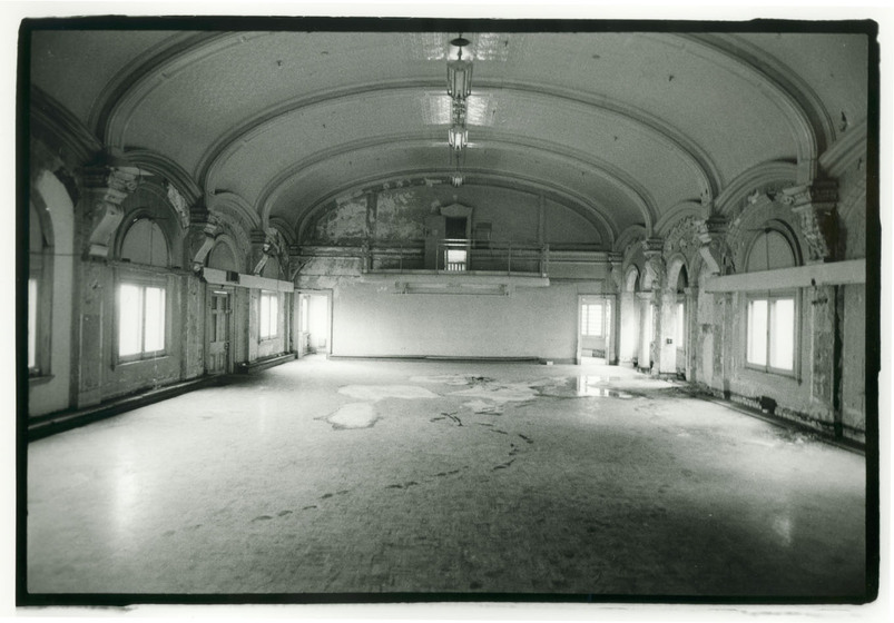 Black and white image of an empty ballroom. Large arched windows line each side of the room, with decorative fixtures surrounding the window frames. There look to be large patches on the floor as though it is in disrepair.