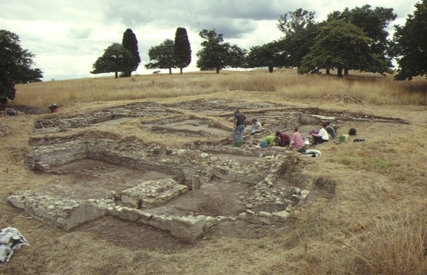 An archaeological dig site with the remains of brick walls sticking out from the ground. In the background a group of people can be seen digging in the site.