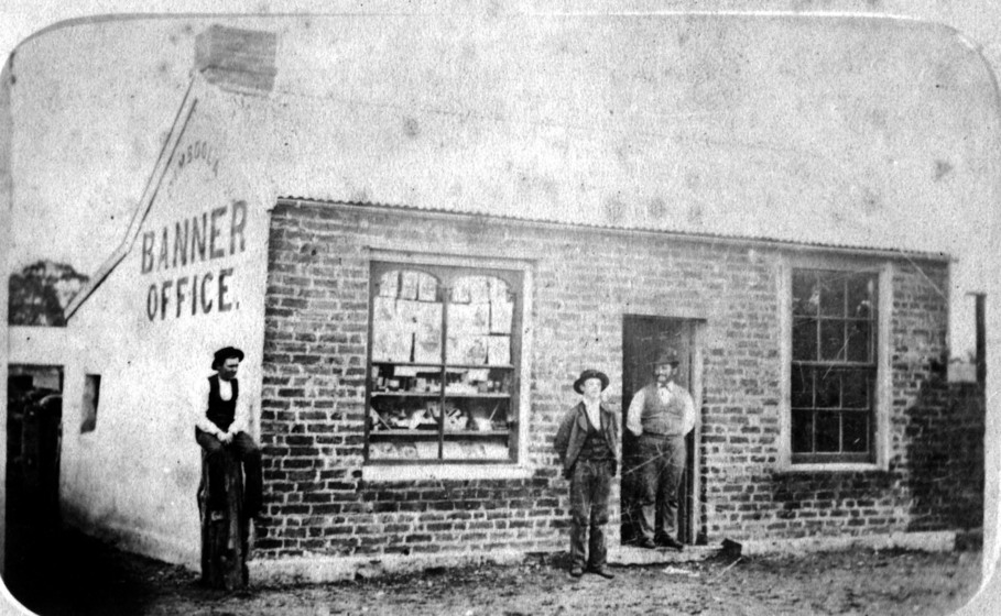 Black and white photograph of three men standing in front of a brick building. On the side of the building a printed sign reads 'Banner Office'.