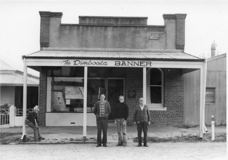 Three men standing in front of a brick shop front, with a tin rood awning strecthing across the footpath. To the left, a younger boy leans on one of the verandah posts.