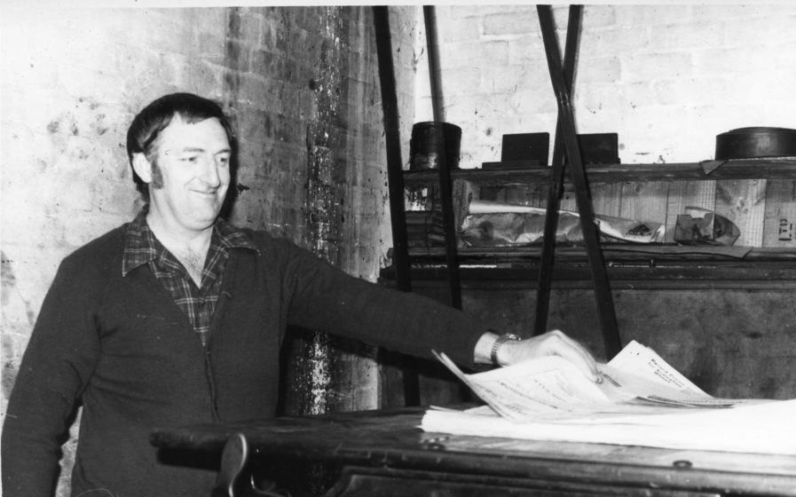 A smiling middle aged man lean over a high table and pulls up a newspaper page from a pile of paper.