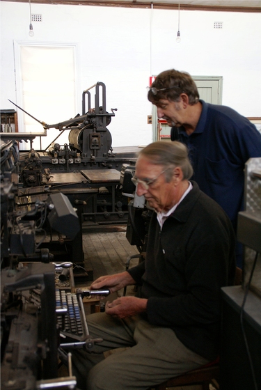 An older man sits at a machine resembling a type writer. Behind him another man stands close by watching the sitting man. Behind them stand various large types of printing press machinery.
