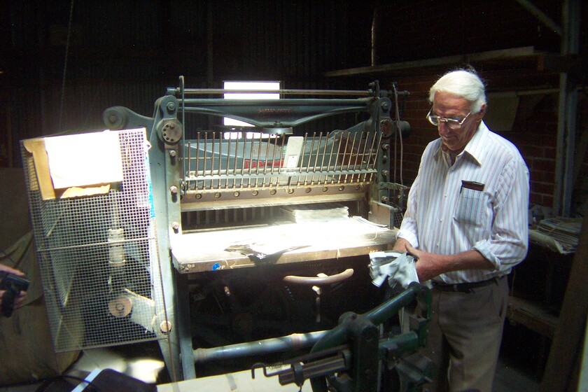 An older man tends to a large piece of machinery inside a brick room. He looks to have a cloth in his hand and is cleaning something.