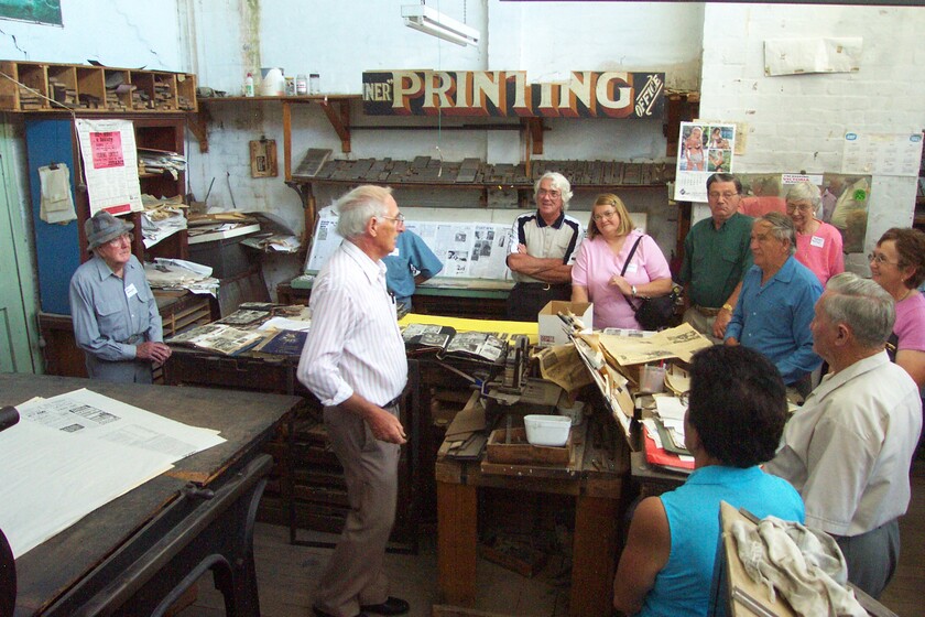 An older man stands in the middle of a room with lots of workbenches and newspapers scattered throughout. He is speaking to a group of about 10 people who are all listening intently.