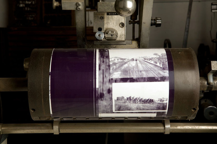 A large metal  and glass cylinder attached to a steel frame. Inside one end of the cylinder you can see black and white photographs pressed up against the glass. In the other end of the cylinder you can see blue ink.