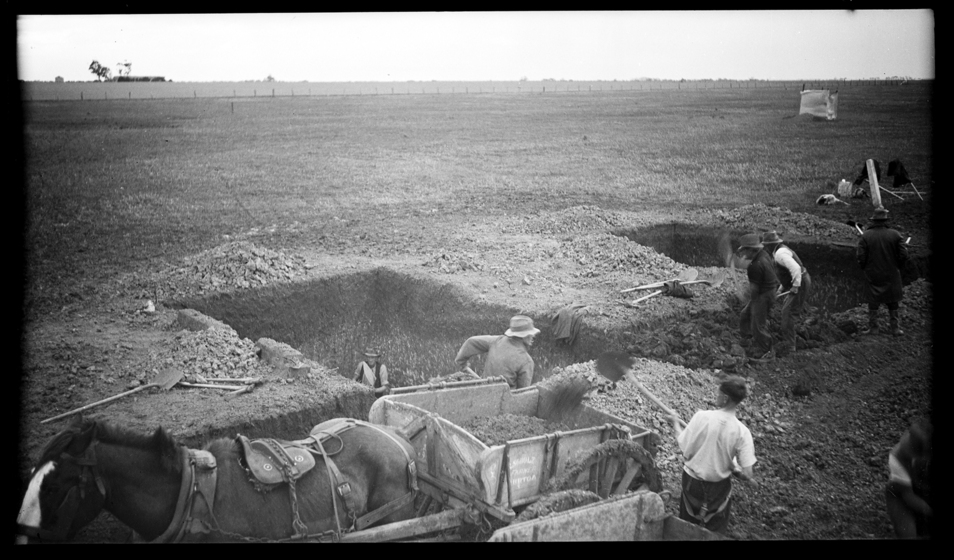 Black and white image of a group of men digging large square holes into the ground. A horse and cart stands to the side, with the cart full of dirt from the holes.