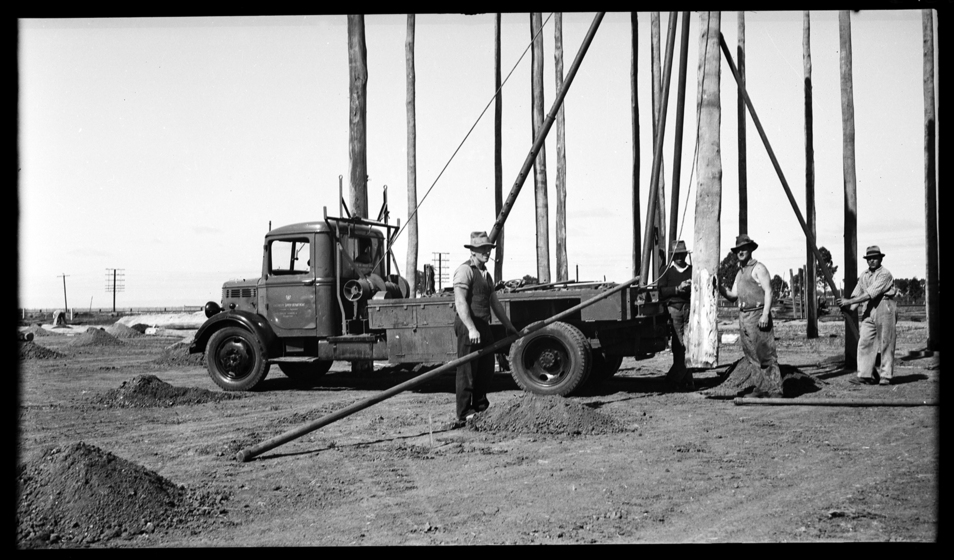 A group of men in working clothes and hats place large wooden poles into holes in the ground. A medium sized truck is positioned between the group of men.