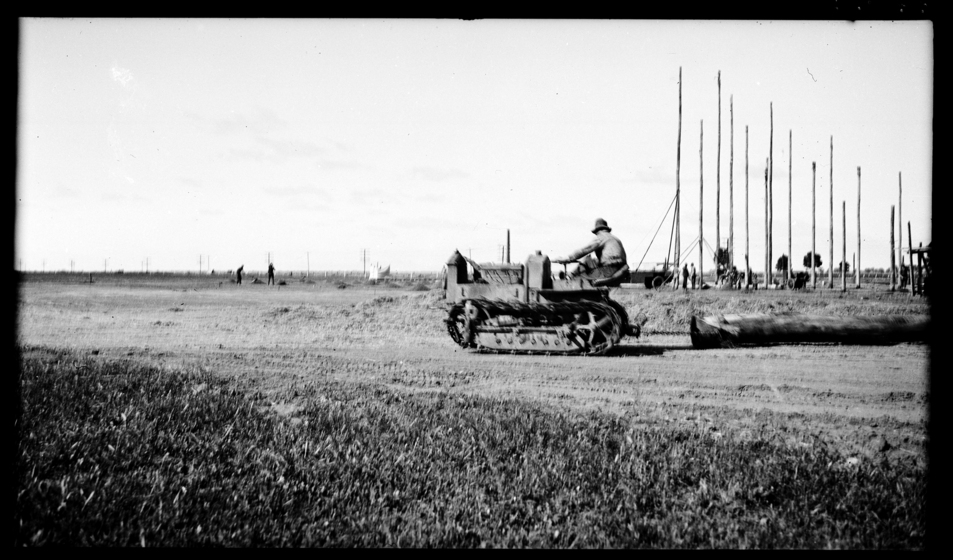 A man sits on a piece of machinary with a large wooden log being dragged behind him. In the distance, upright poles are standing in the ground.