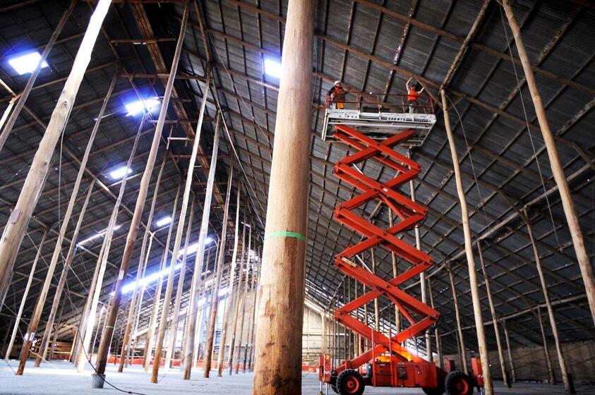 The interior of a large, tall shed. The roof is being held up by a large number of wooden stick poles. A scissor lift is raised to the roof, with two men working inside of it.