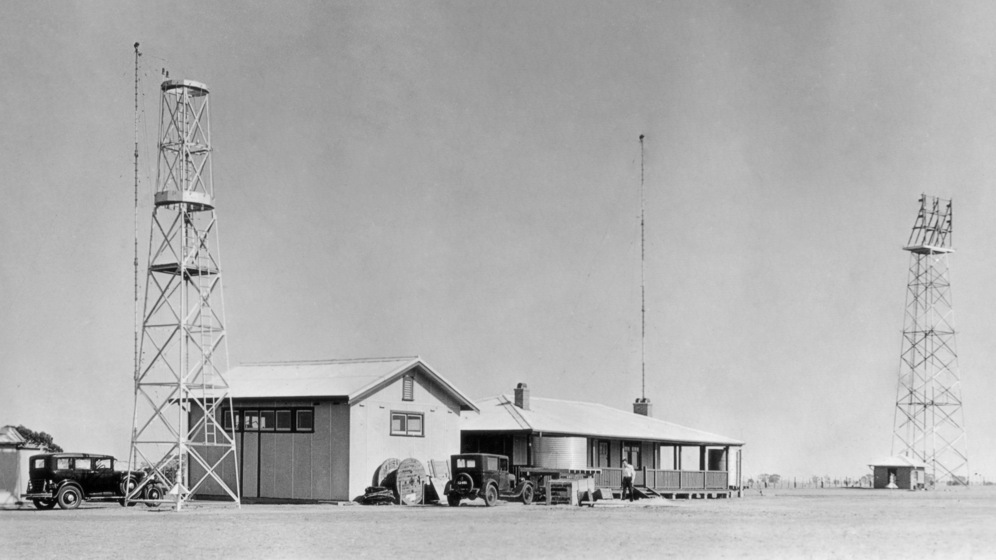 Black and white image of two weatherboard buildings - one with a verandah and tank, and one without. To either side of the buildings are two tall metal radio masts, as well as a single antenna coming off one of the houses.