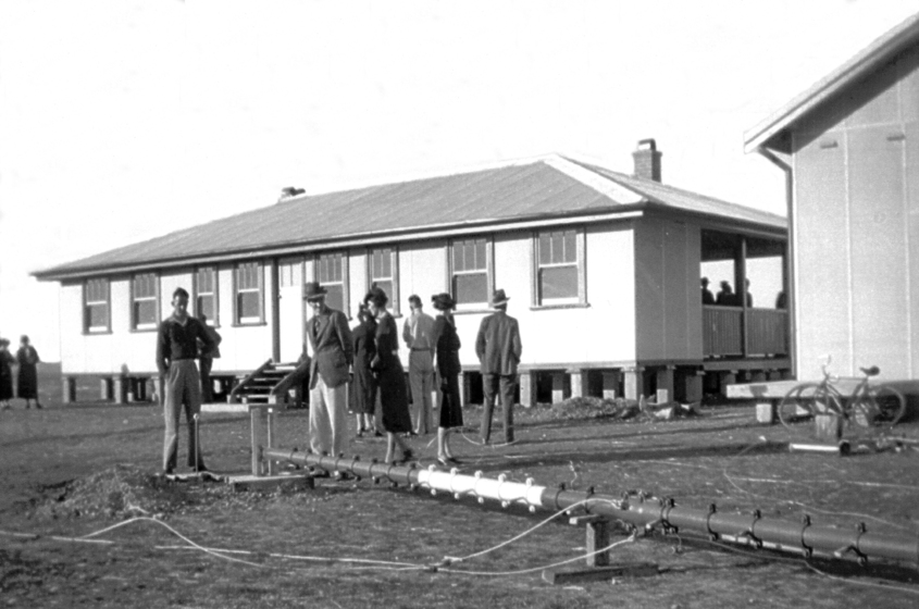 A group of well dressed people stand on a grass patch in front of a weatherboard building. On the ground lays a large metal radio mast