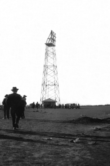 A tall metal radio beacon stands in an open field. Scattered around the base are well dressed people admiring the radio beacon.
