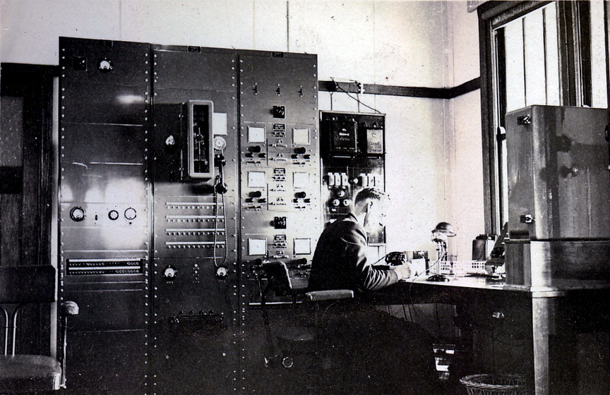 A man sits at a desk, poised to write something down. On the wall behind him are a large number of switches, knobs and equipment.