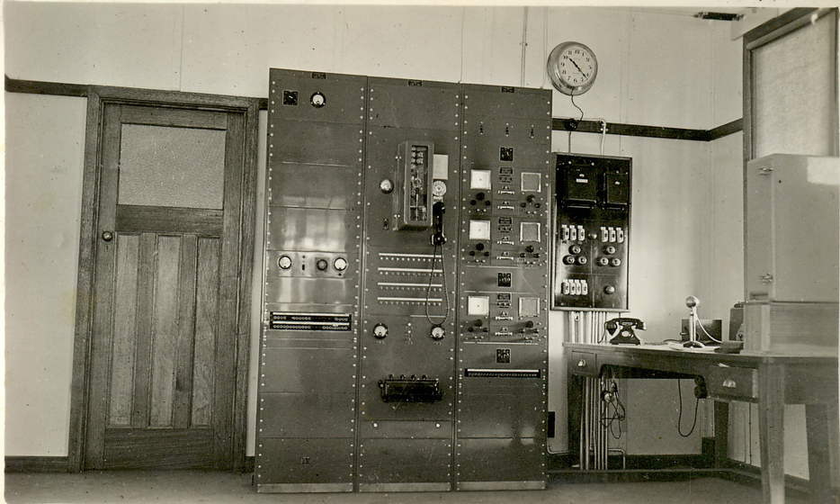 Black and white image of a room containing a large cupboard with knobs, switches and meters on the front of it. To the right of the equipment, a switchboard can be seen on the wall, and a nearby table holds a microphone and telephone.