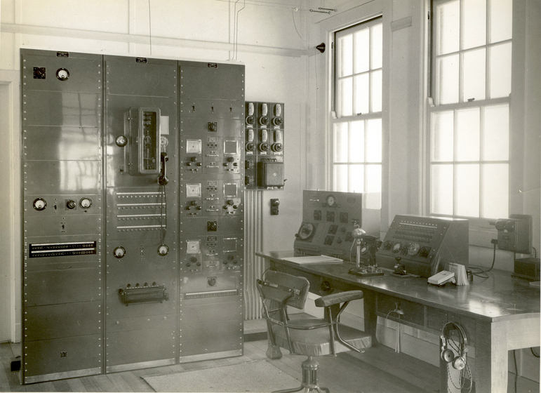 Black and white image of a room containing a large cupboard with knobs, switches and meters on the front of it. To the right of the equipment, a switchboard can be seen on the wall, and a nearby table holds a microphone and further equipment with dials and knobs. A headset hangs off the side of the table.