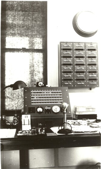 Black and white image of an operator's console on a desk. A microphone stands in front of it, along with a lamp and a desk calendar.