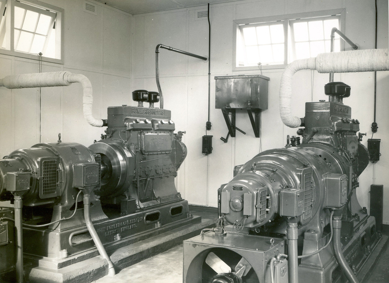 Two large generators stand in a room. They have pipes and cords exiting the generator which attach to the interior walls.