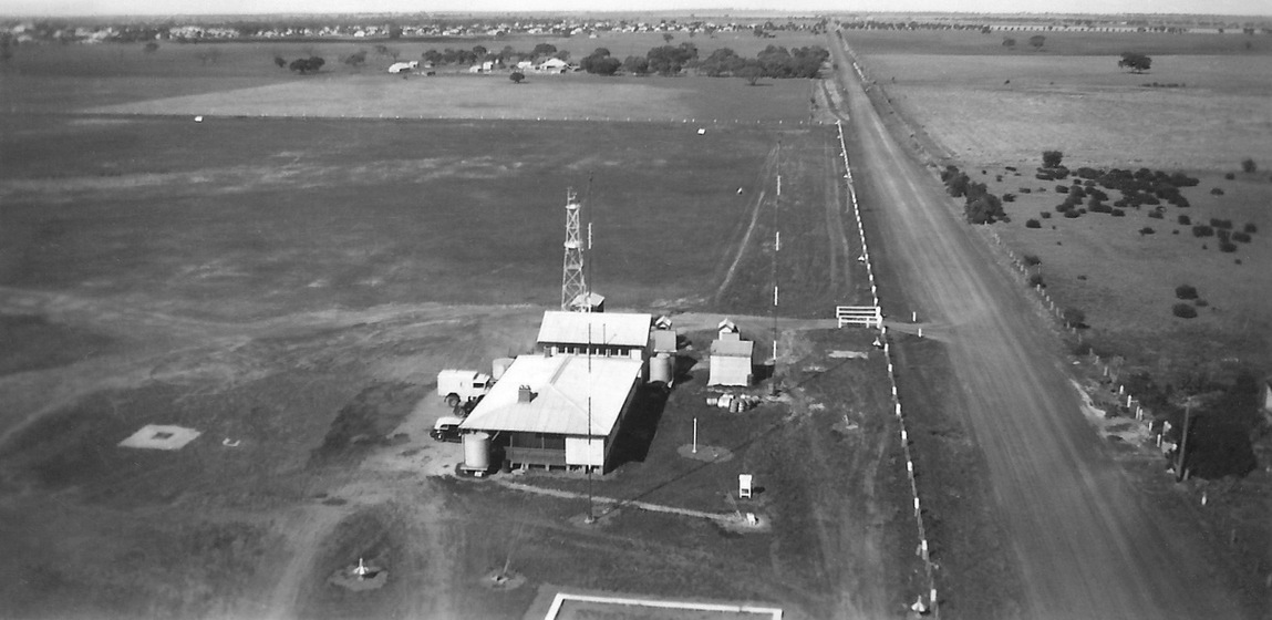 Aerial view of a building standing next to a dirt airstrip. Tall radio antenna's come off the roof of the building.