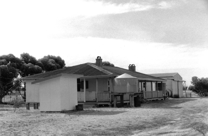 A weatherboard building on stumps stands in an open grassed space. It has a wrap around vernadah on one side of the building, and a raised water tank out the front. A shed can be seen in the distance and in the foreground is a small outhouse with no windows.