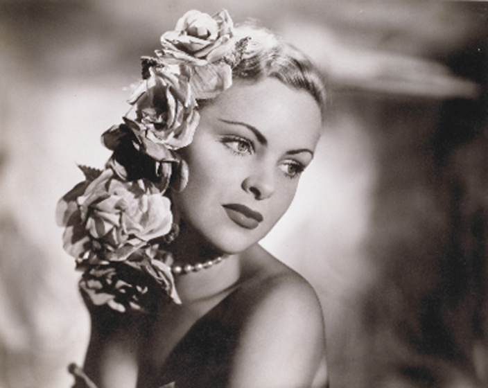 A model poses for the camera with her head tilted slightly to the side. She has paper flowers tied into her hair and a pear necklace around her neck.