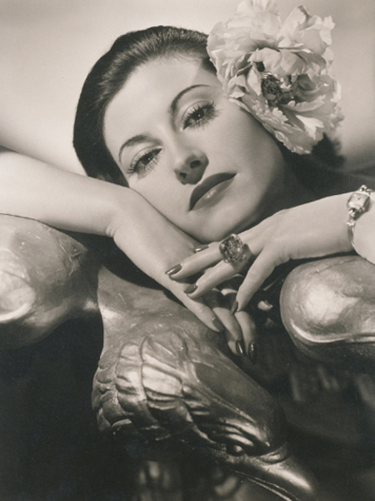 A model leans on a bronze sculpture of a bird. She is wearing decorative costume jewelry and has a large paper flower attached to the side of her head.