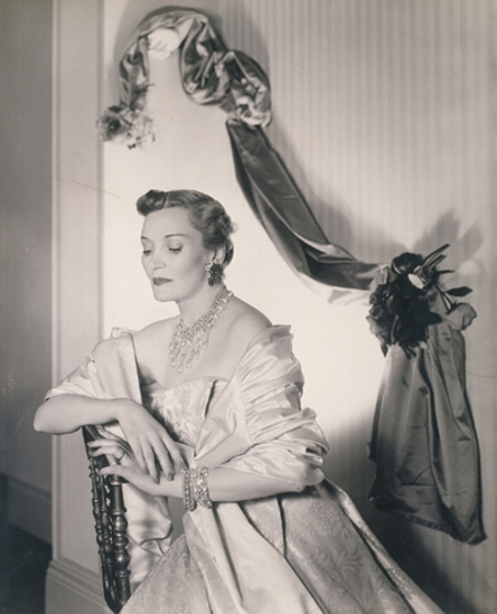 A model sits on a chair, leaning one of her arms on the back of the chair. She is dressed in a ball gown and has elaborate costume jewelry on her wrists, neck and ears. Behind her on the wall is a satin sheet, bunched in positions with flowers to form a decorative backdrop.