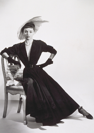A woman sits on the edge of a chair, one arm leant on the back of the chair and the other with her hand on her hip. She is pointing her leg out to the side. She is wearing a floor length black structured dress, black gloves and a large white feathered hat.