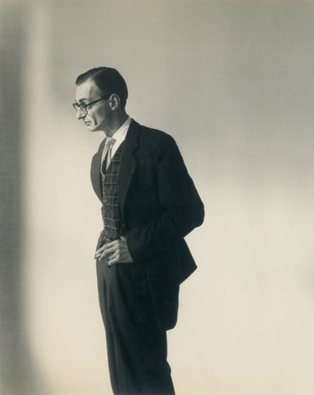 A man stands with his side facing the camera. He is dressed in a suit and vest, with black rimmed glasses. One hand is holding a cigarette and the other is in his pocket.