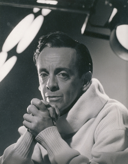 An older man stares straight at the camera. His hands are clasped together and he is resting his head gently on them. He is wearing a white turtle neck sweater.