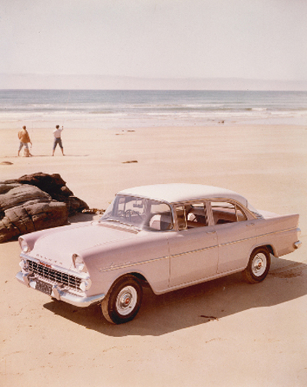 A 1950s model holden, pink in colour, is parked on the beach. Behind the car are two men standing looking out to sea.