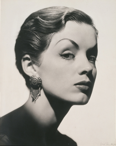 A black and white portrait of a woman's face. She is wearing detailed makeup and one dangling earring.