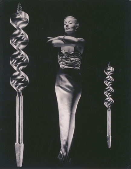 A black and white image of a woman standing upright, posing with her legs tight together and her arms crossed over one another like a genie stance. To either side are sculptures that resemble drill parts.