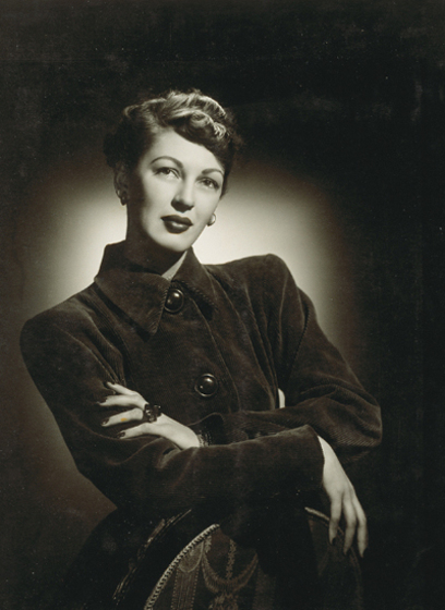 A woman poses for the camera. One arm is draped over the back of a chair, whilst the other lies across her body. She is wearing a buttoned down coat with a large triangular collar.