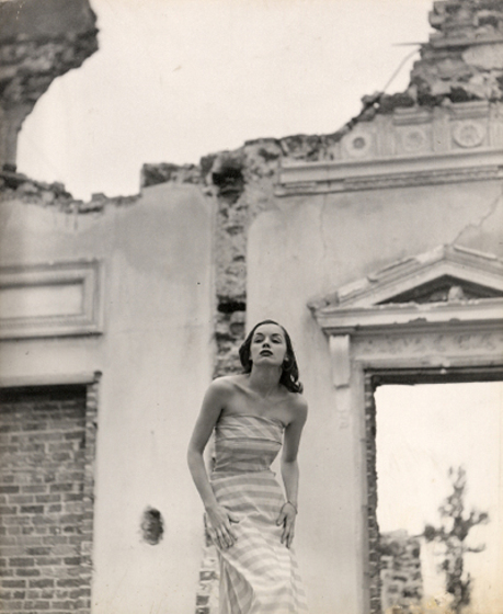 A model stands slightly hunched over, posing in front of the facade of a crumbling old building.