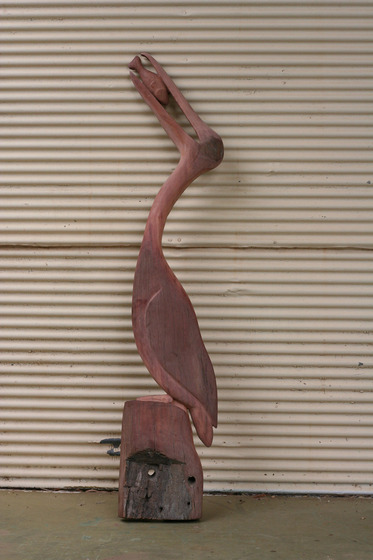 Wooden sculpture of a pelican sitting on a log or rock. His bill is up in the air and he is holding a fish in his mouth.