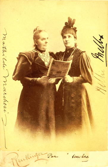 A sepia photograph of two women dressed in full length dresses, standing close to each other and both holding the same piece of paper in their hands.
