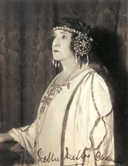 A woman dressed in a decorative silk dress stares into the distance, not looking at the camera. She is wearing a decorative head piece that features pearls.