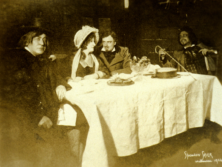 A sepia photograph of a group of four actors sitting around a table. All of the actors are in costume, including a man in a top hat, and a woman in a bonnet.