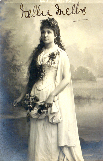 A woman in a full length gown stands looking slightly to the left of the camera. Her dress is decorated with floral emblems, and she is holding fabric, most likely attached to her dress, in both hands. She is wearing flowers in her hair, and her long hair falls over her shoulder on one side.