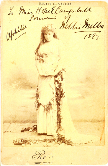 Sepia full length photograph of a woman standing, looking down at the ground and holding her hands together in front of her waist. She is wearing a floor length gown that is decorated with floral garlands that are draped across the dress and spread onto the ground.