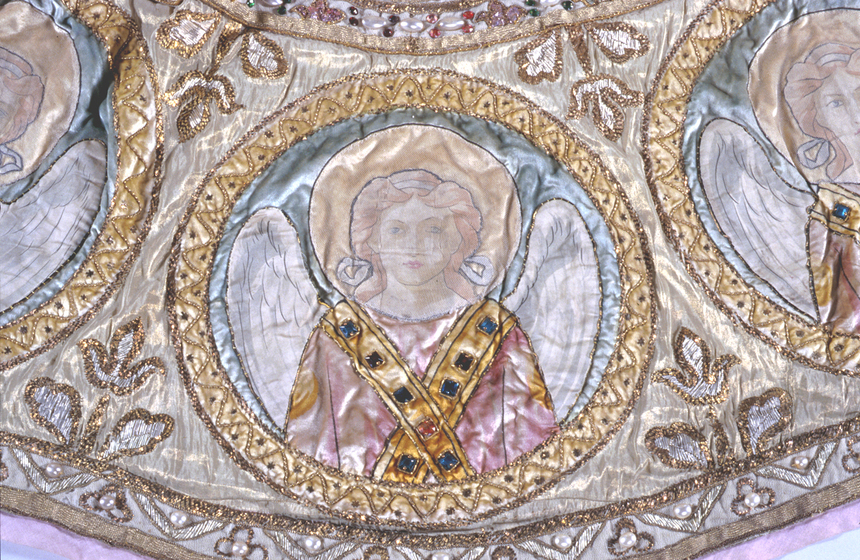 Detailed view of a segment of a gold cloak. An embroidered silk angel, featuring jewels, is surrounded by a gold beaded circle. Gold thread leaves frame the circled angel.